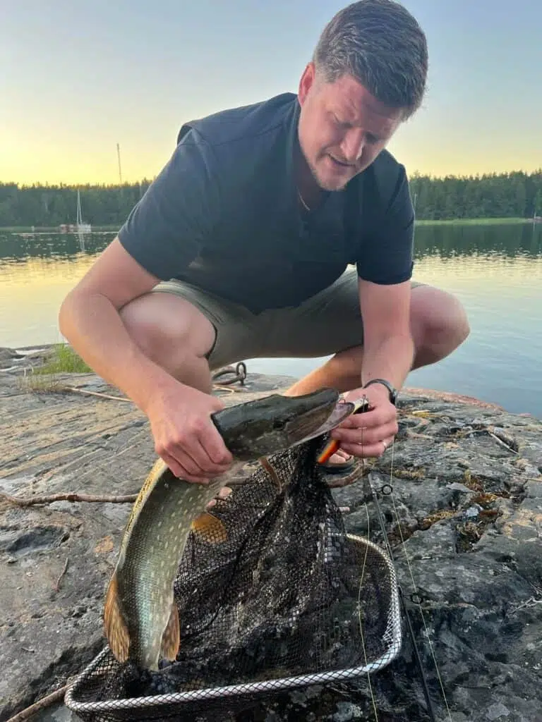 En person som håller i en fisk utomhus vid en sjö använder en svart kylväst