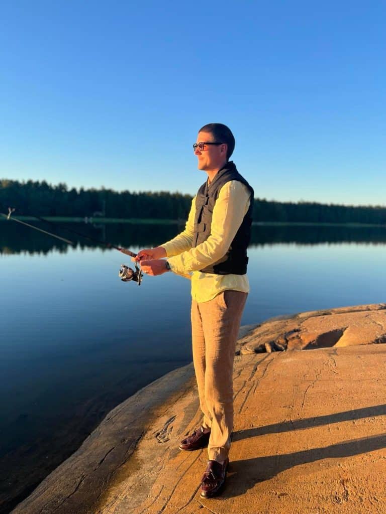 a man fishing standing on rocks next to a lake. He wears a black cooling vest
