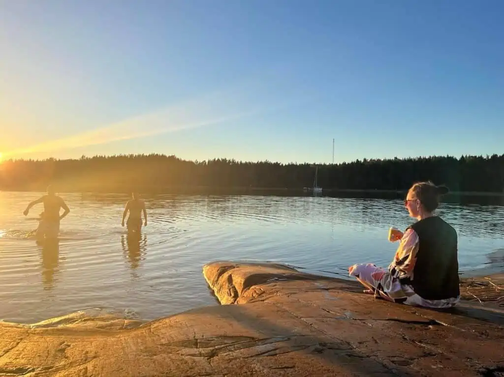 En tjej sitter på klipphäll iklädd en svart kylväst och tittar ut mot vänner som badar en solig sommarkväll