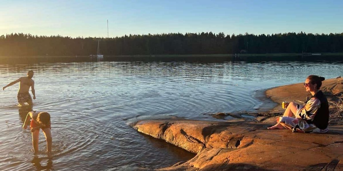 En tjej sitter på klippblock en solig sommarkväll vid vattnet. Hon tittar ut mot två personer som badar. Hon sitter iklädd en svart kylväst